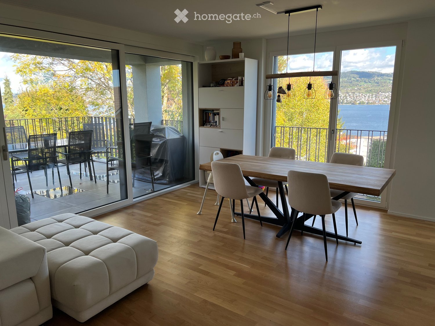 Modern living and dining area with hardwood floors, large windows offering a scenic view, a sofa, a dining table with chairs, and a shelf with decorative items.