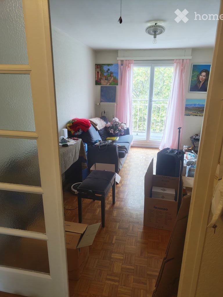 A furnished living room with a blue sofa, wooden floor, a small table, a stool, and a balcony with pink curtains. There are paintings on the walls and some boxes and luggage in the room.