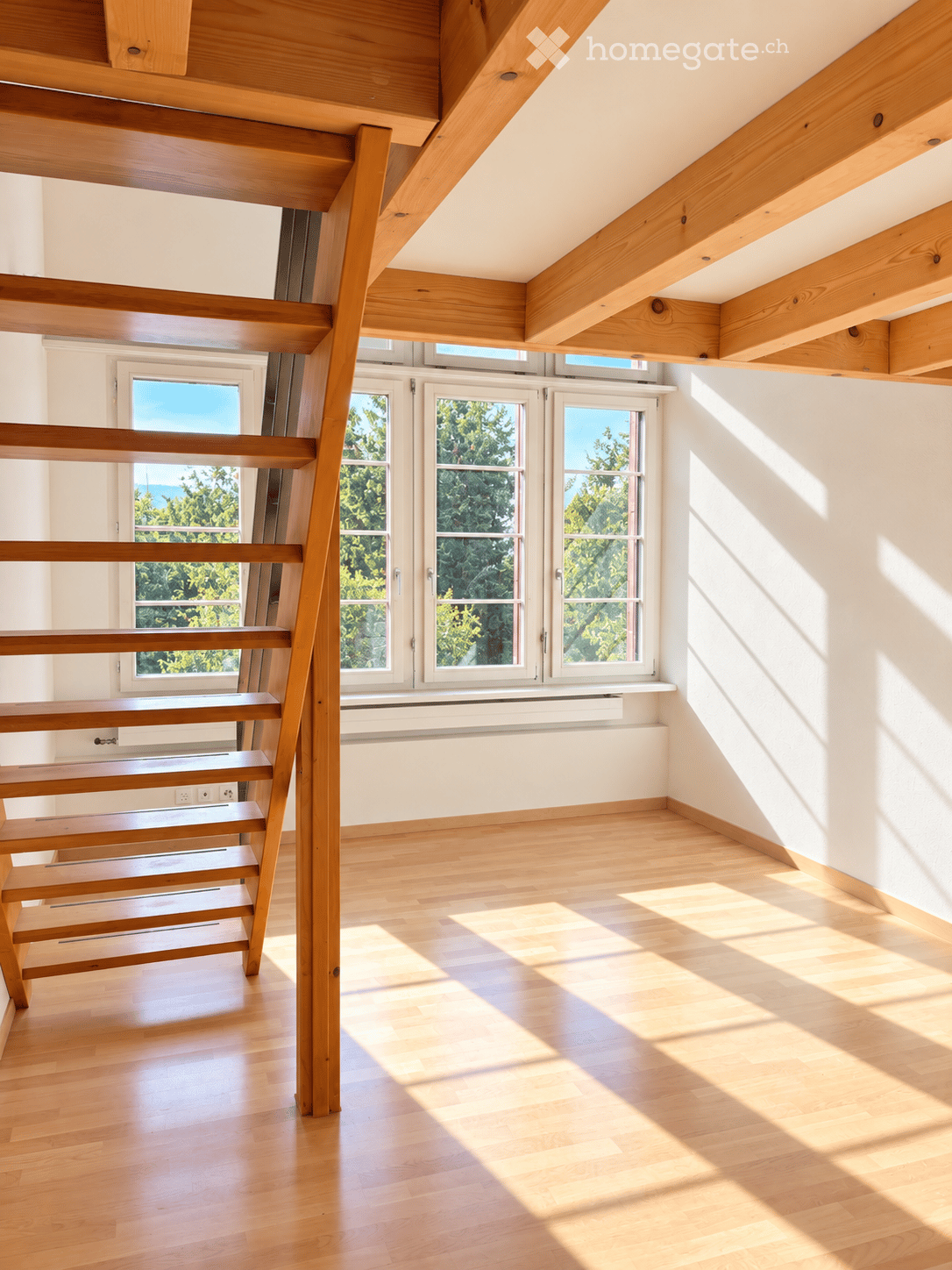 Bright, empty room with wooden flooring and exposed wooden beams on the ceiling. Large windows allow natural light to fill the space, casting shadows on the floor.