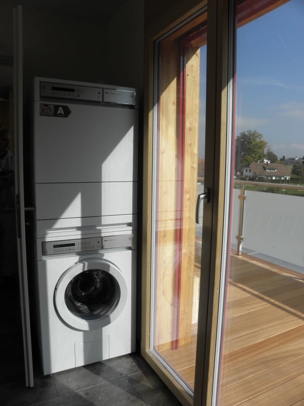Modern bathroom with a washing machine and dishwasher stacked vertically, large window with view, wooden floor, and tiled walls.