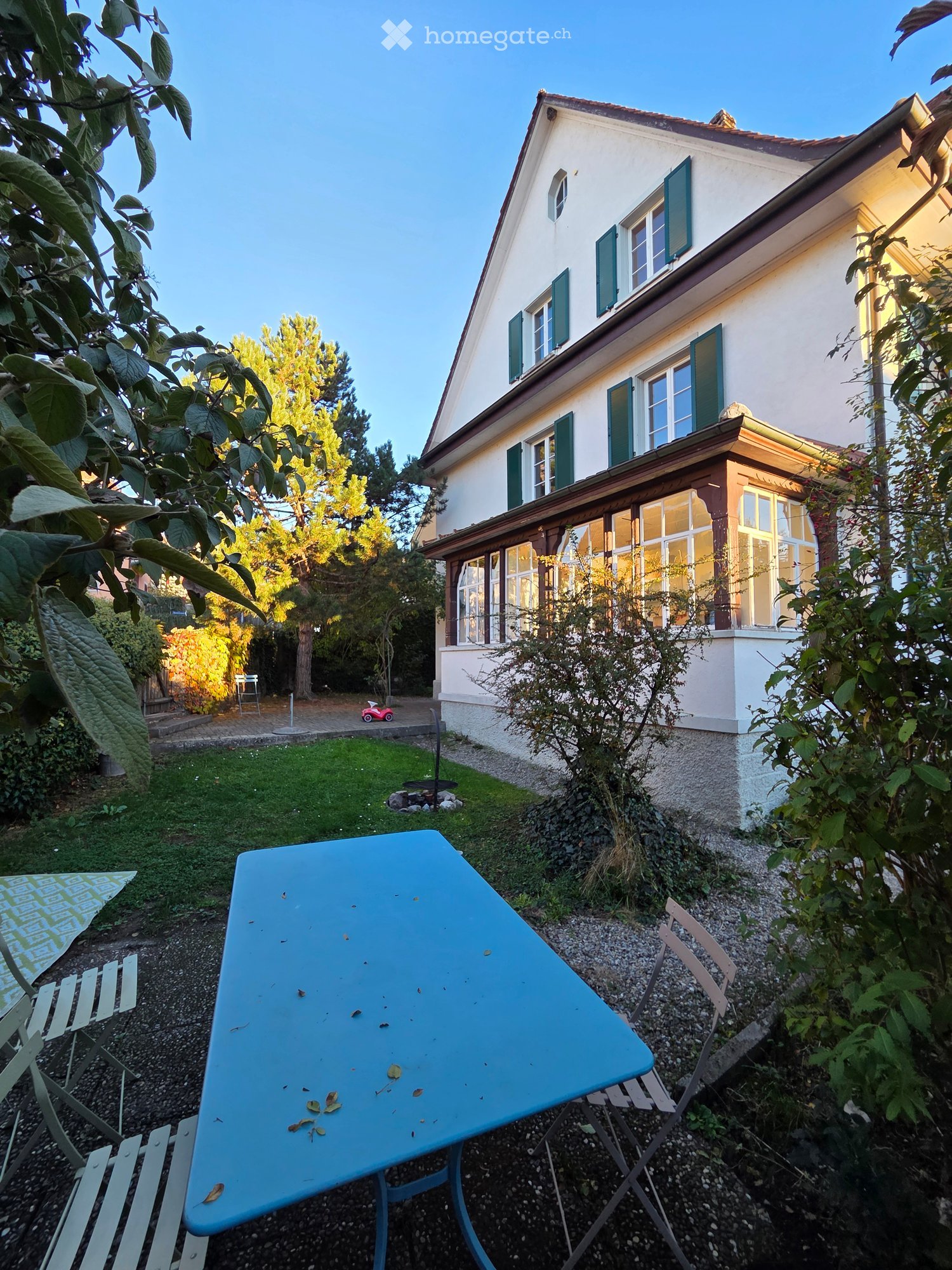 Garden area with a blue table and chairs, a small lawn, and a two-story house with green shutters and large windows in the background.