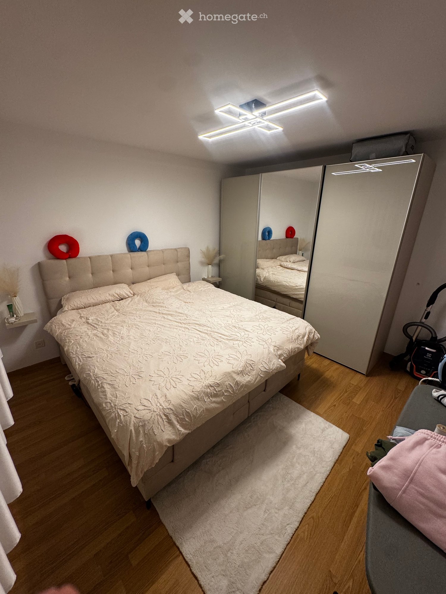 Modern bedroom with a large bed, mirrored wardrobe, nightstand, and vacuum cleaner in the corner.
