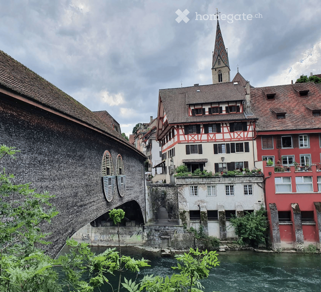 A historic European town scene with traditional buildings, a church spire, and a canal running through the area.