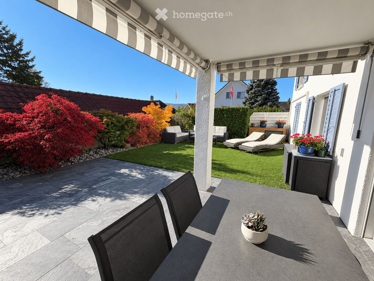 A modern balcony with a grey stone floor, a table with two black chairs, and a small potted plant. The balcony has a striped awning and overlooks a well-maintained garden with lounge chairs, a small table, and colorful plants.