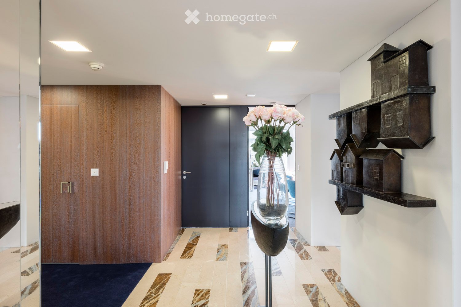Modern hallway with polished marble tiles, wooden doors, recessed lighting, and decorative shelves with miniature houses. A vase with pink roses is placed on a sleek black pedestal table.