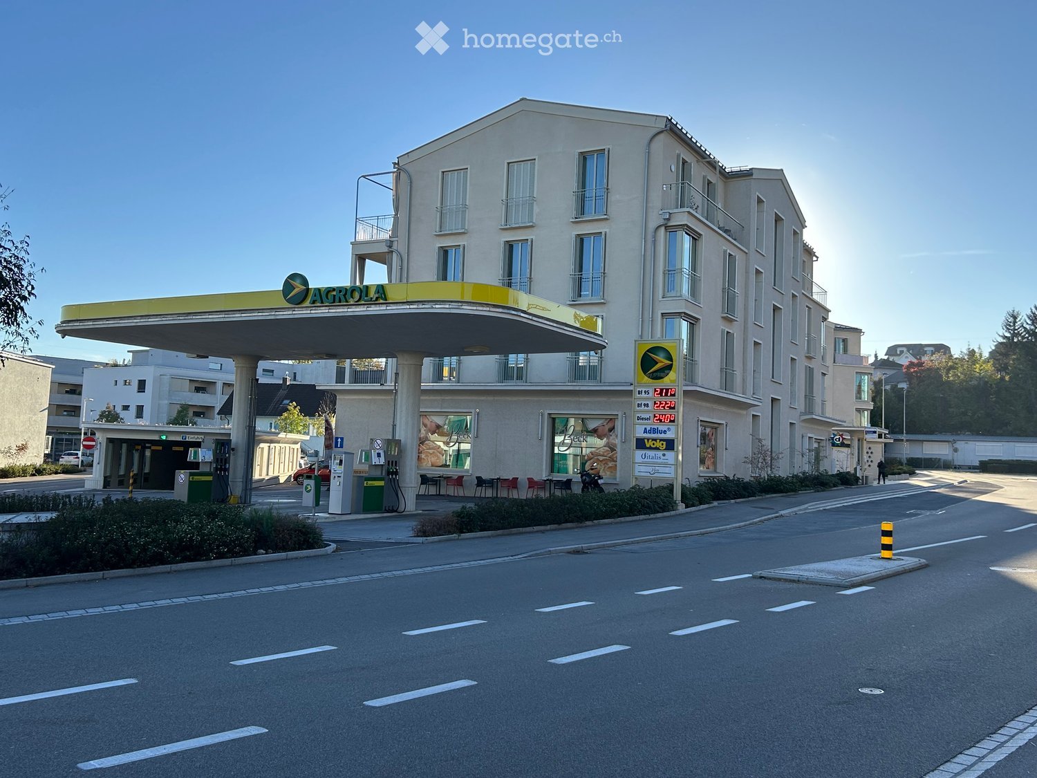 Multi-story building with a modern facade, featuring balconies and large windows. A gas station with a prominent 'AGROLA' sign is attached to the building.