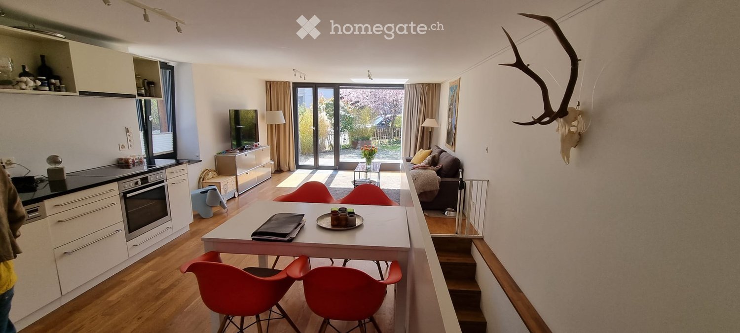 Open-plan living and dining area with a sofa, coffee table, dining table with chairs, kitchen area with oven and fridge, and decorative skull on the wall.