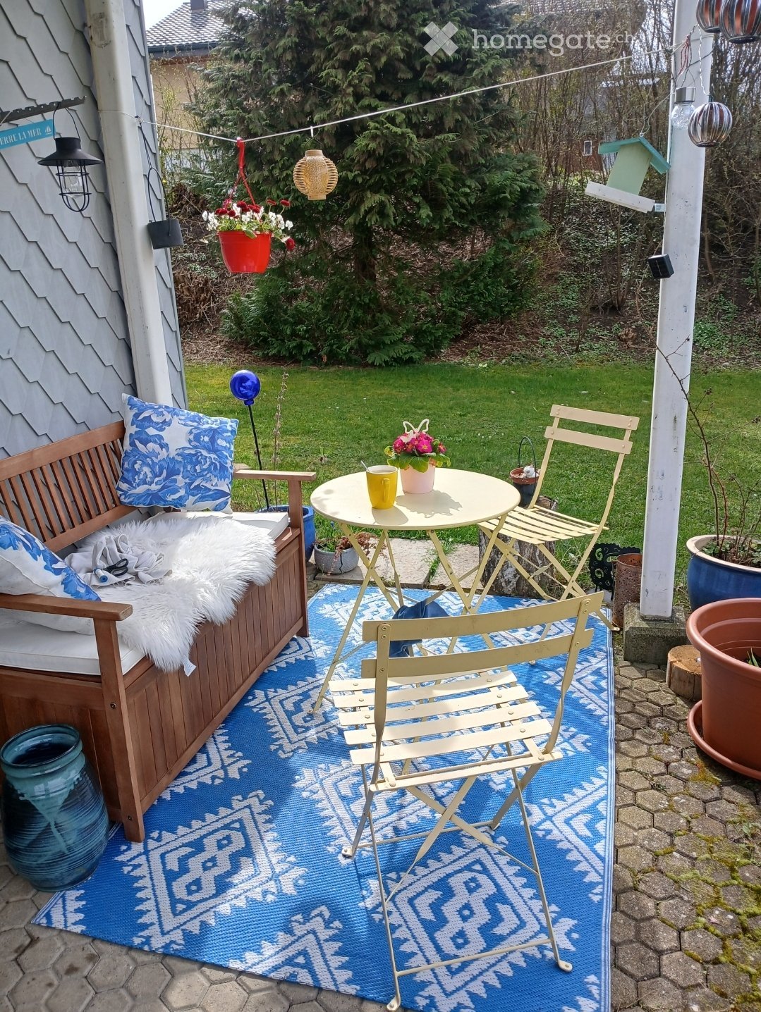 A cozy outdoor seating area with a wooden bench, a small round table, two folding chairs, and a blue patterned rug. There are potted plants and hanging decorations, with a well-maintained lawn in the background.