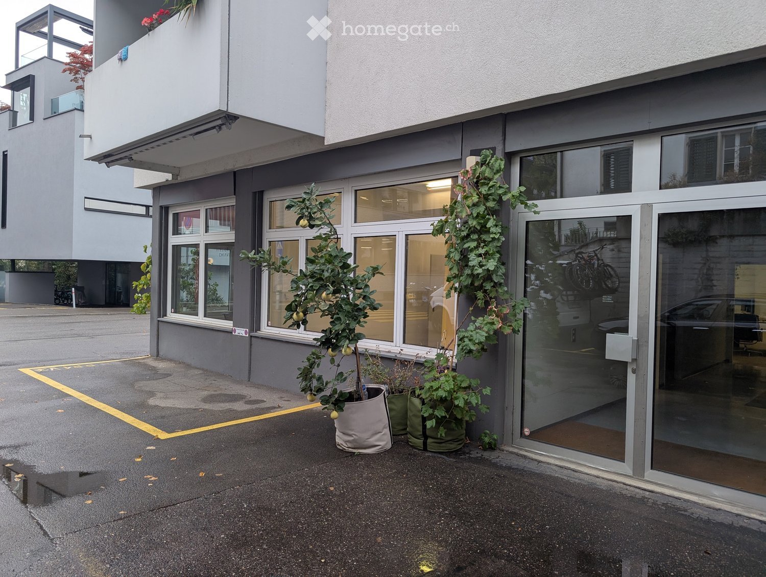 Modern building entrance with glass doors, potted plants, and a small parking area.
