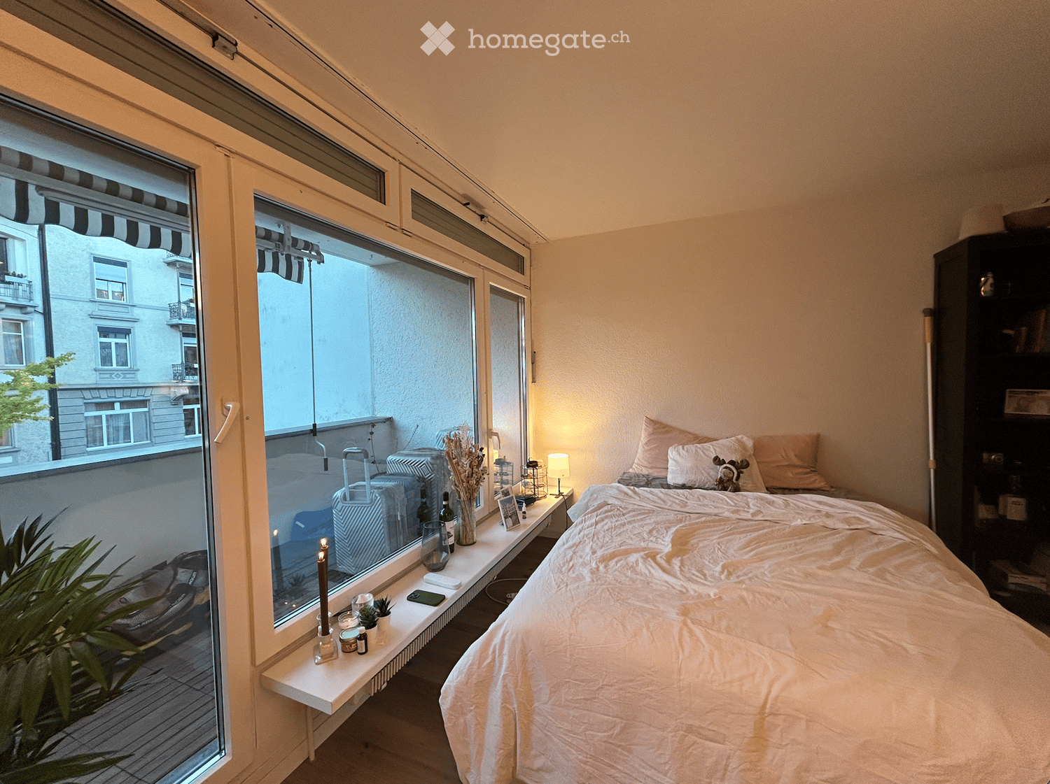 Cozy bedroom with a large window offering a view of the street, a neatly made bed with pillows and a stuffed animal, a nightstand with a lamp, decorative items, and a suitcase visible through the window.