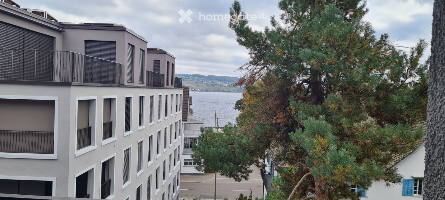 Modern multi-story building with balconies and large windows, overlooking a body of water with trees in the foreground.