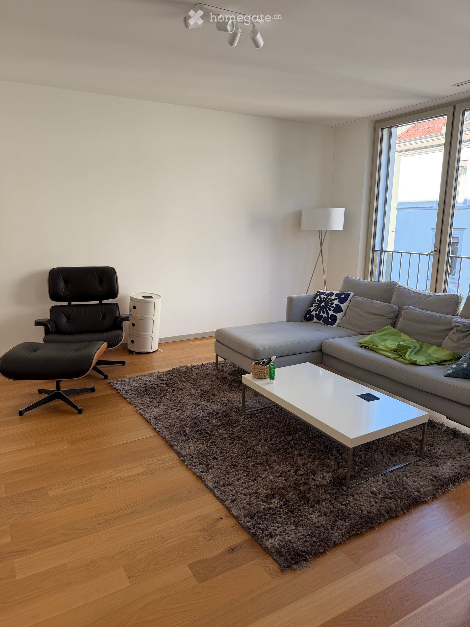 Modern living room with a grey sectional sofa, white coffee table, black leather chair, floor lamp, and a shaggy rug on a wooden floor.