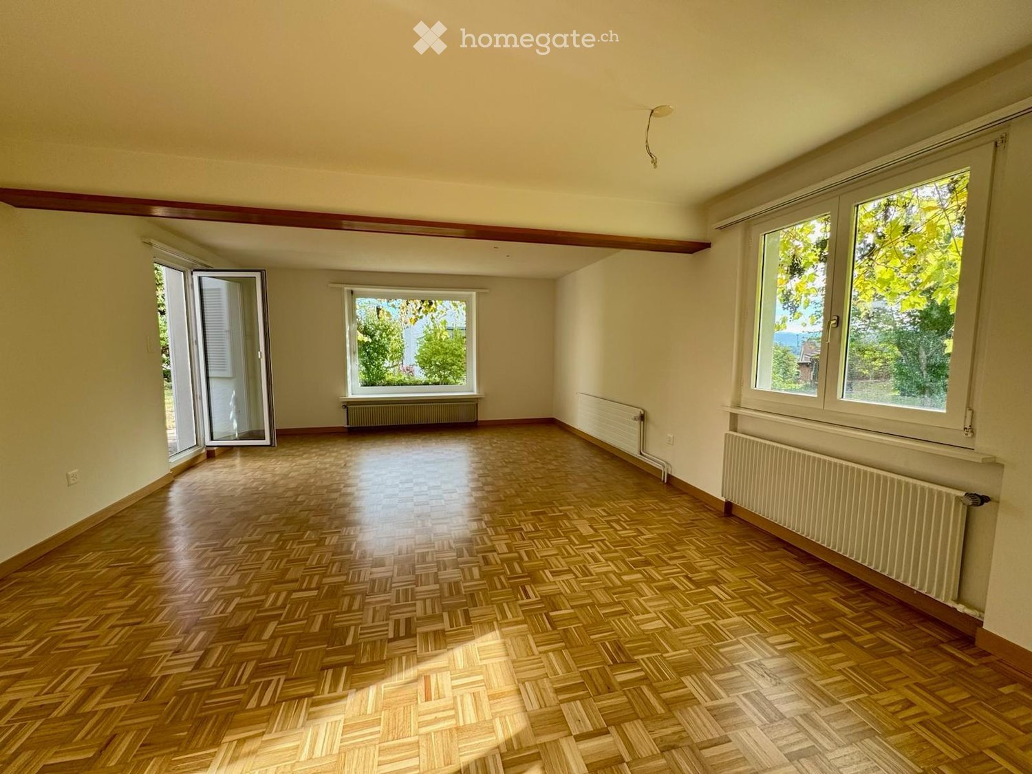 Empty room with wooden herringbone flooring, large windows, radiators, and a sliding glass door leading outside.