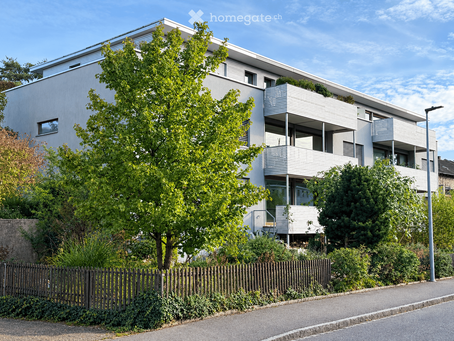 Modern two-story residential building with white facade, large windows, and balconies, surrounded by greenery and a wooden fence.