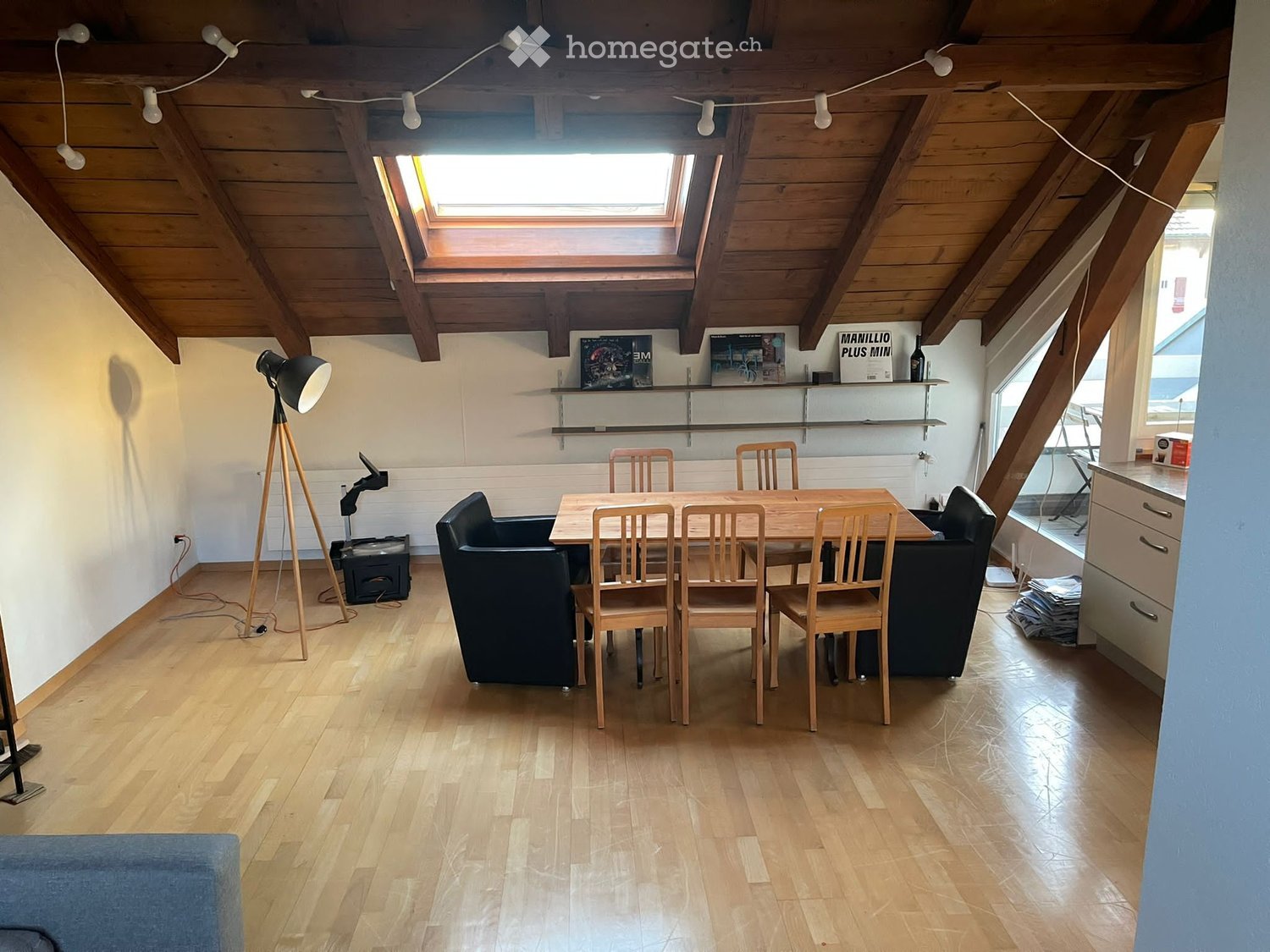 Bright, wooden-floored room with exposed wooden beams and skylights. Features a sofa, dining table with chairs, a floor lamp, and a shelf with books and decorative items.