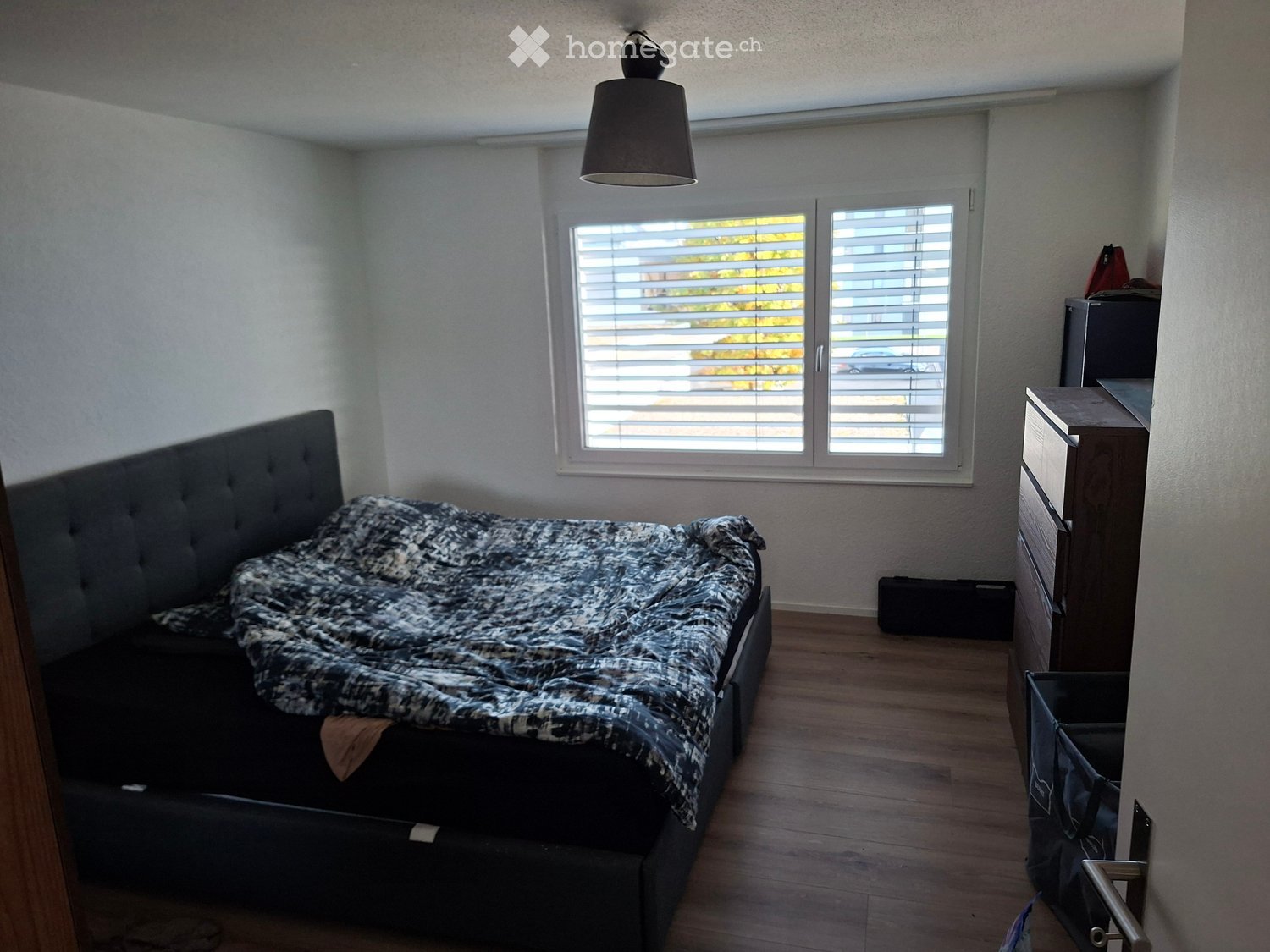 A modern bedroom with a large bed, patterned bedspread, wooden floor, and large windows with blinds. Includes a nightstand, lamp, and a drawer unit.