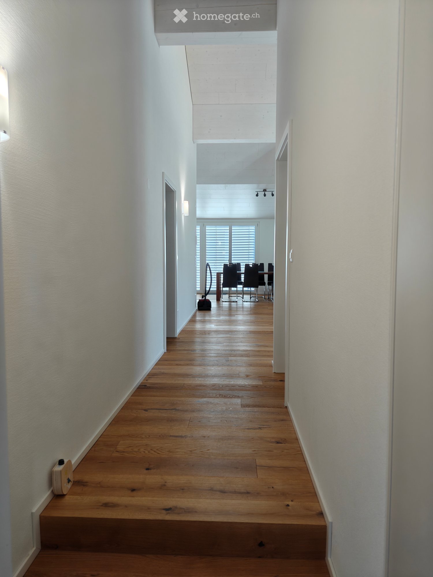 A long, narrow hallway with wooden flooring and white walls, leading to a dining area with chairs and a table visible at the end.