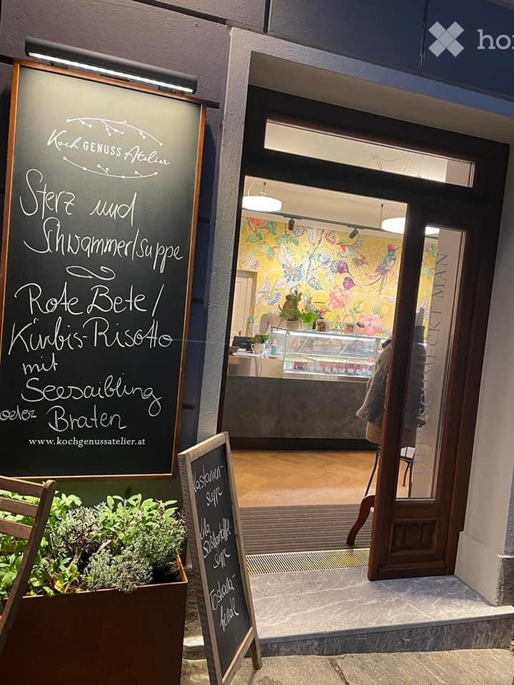 The image shows the entrance to a restaurant or cafe with a chalkboard menu displayed outside. The interior features a colorful floral wall, a counter with various items, and a small seating area visible through the glass door.