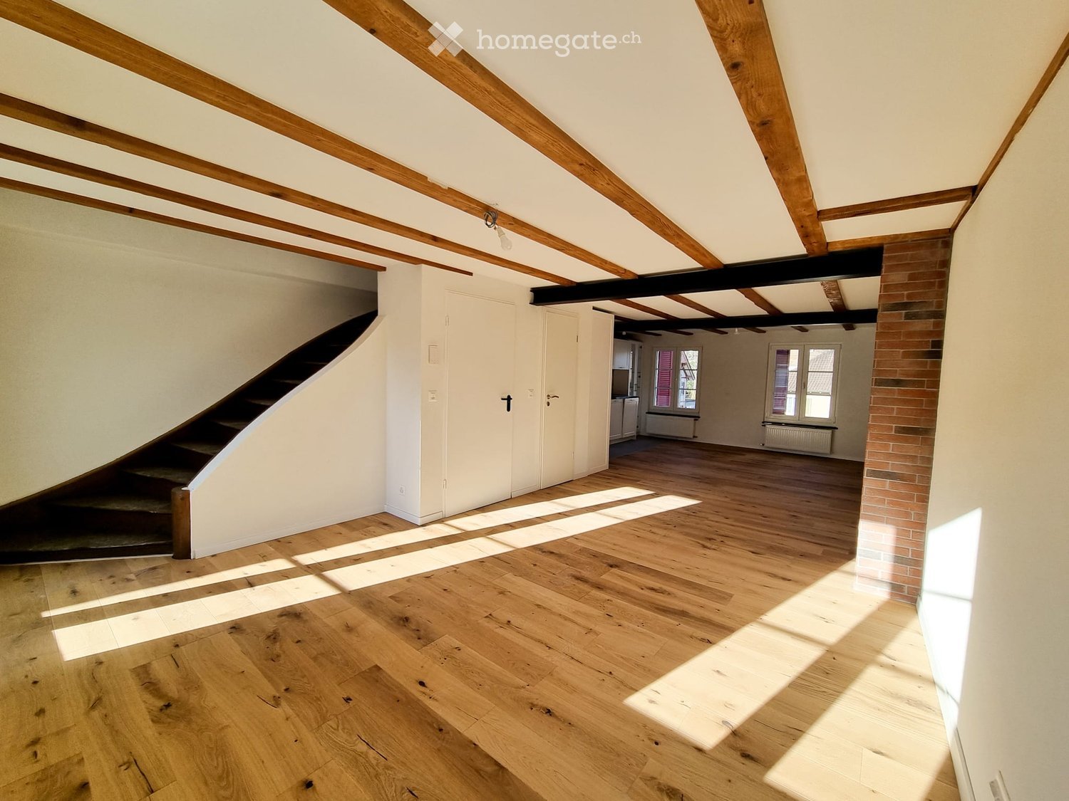 Empty attic space with wooden floors, exposed wooden beams, and natural light coming through windows. There is a staircase leading down, and a brick wall is visible on one side.
