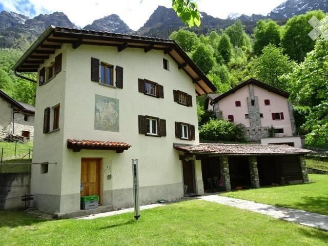 Two-story house with a cream-colored facade, wooden shutters, and a tiled roof, situated in a lush green area with mountain views in the background.