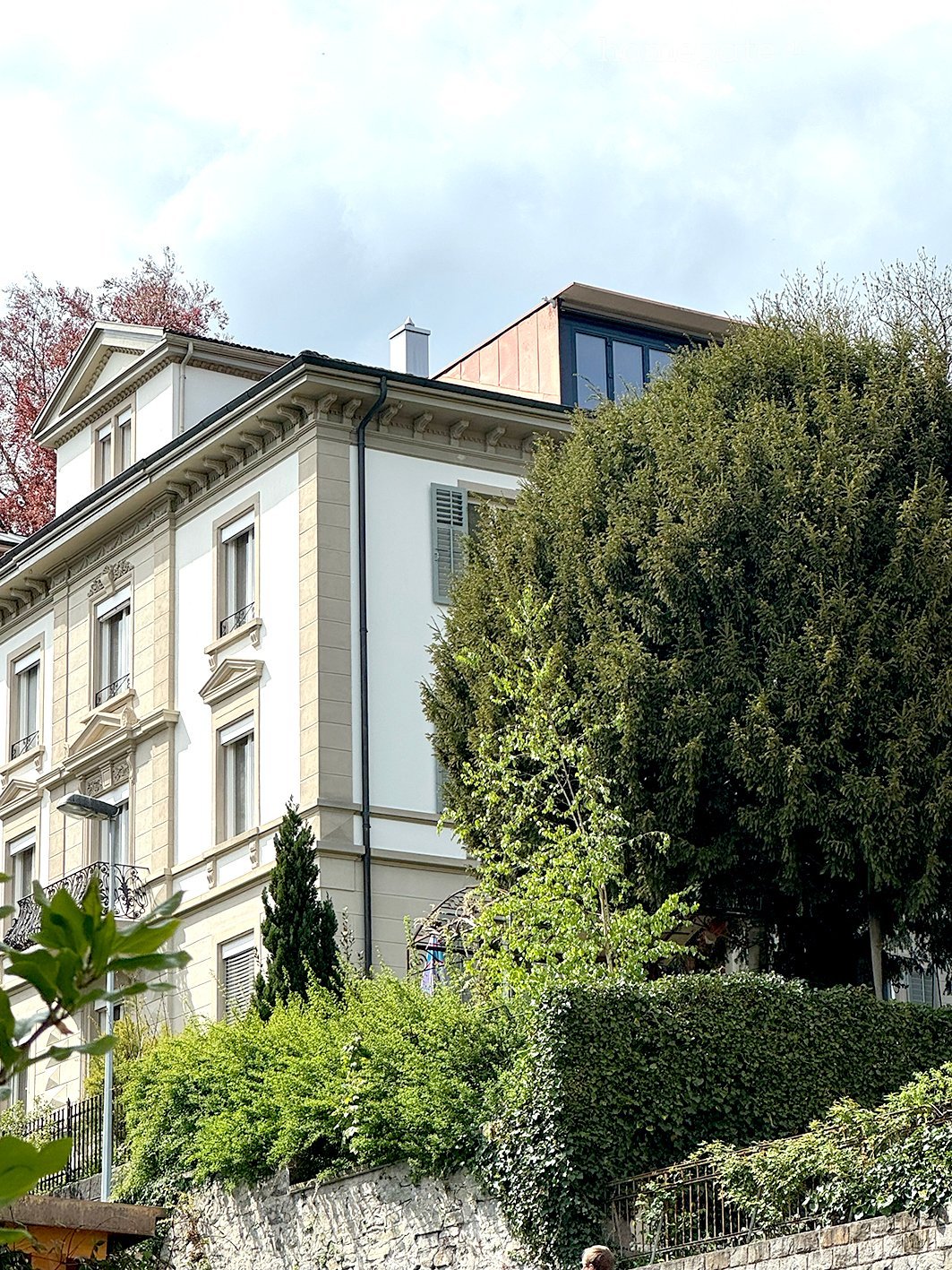 A multi-story residential building with classic architecture, large windows, and decorative stonework, surrounded by lush greenery and trees.