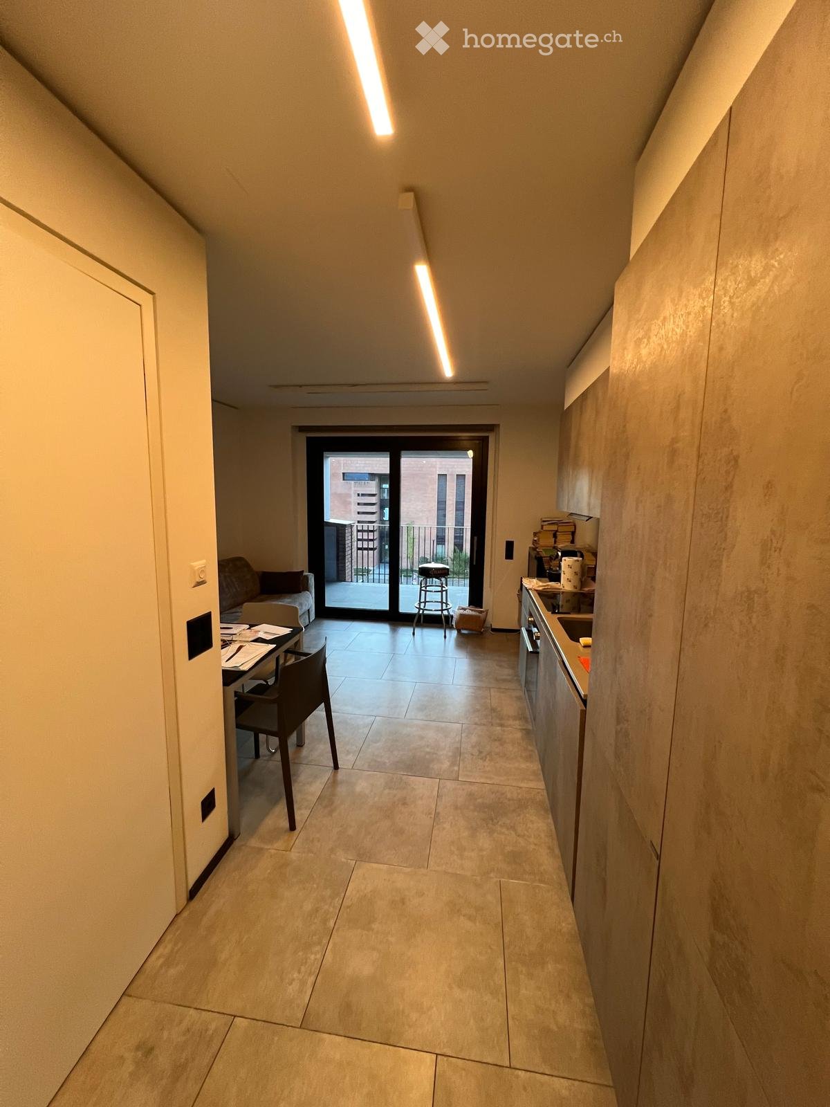 Open-plan living and dining area with a kitchen on the right, a sofa on the left, and a balcony accessible through large glass doors.