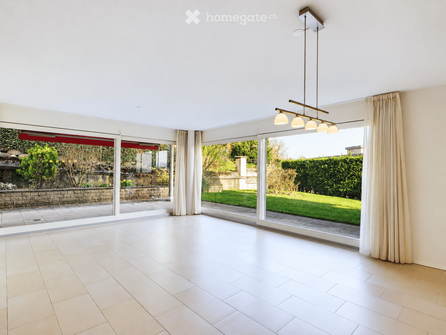 Bright, empty room with large glass sliding doors leading to a garden, white tiled floor, and modern pendant lighting fixture.