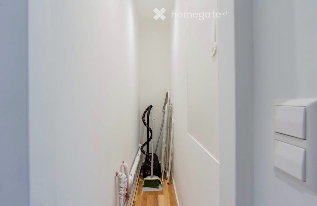 Narrow hallway with light wooden flooring, white walls, and cleaning supplies stored in the corner.