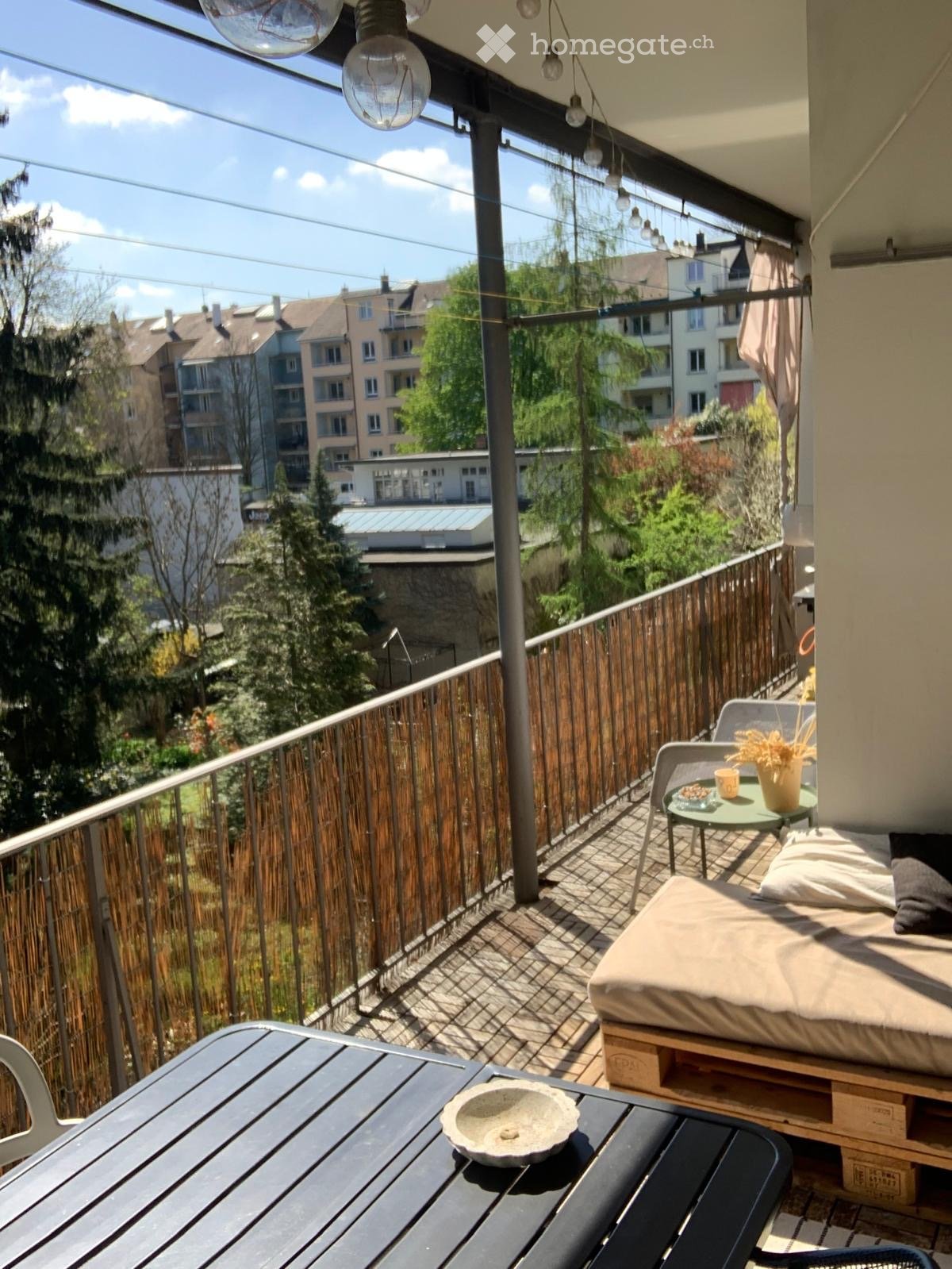 Balcony with wooden decking, bamboo privacy screen, daybed, small table, and chairs, overlooking a cityscape with trees and buildings.