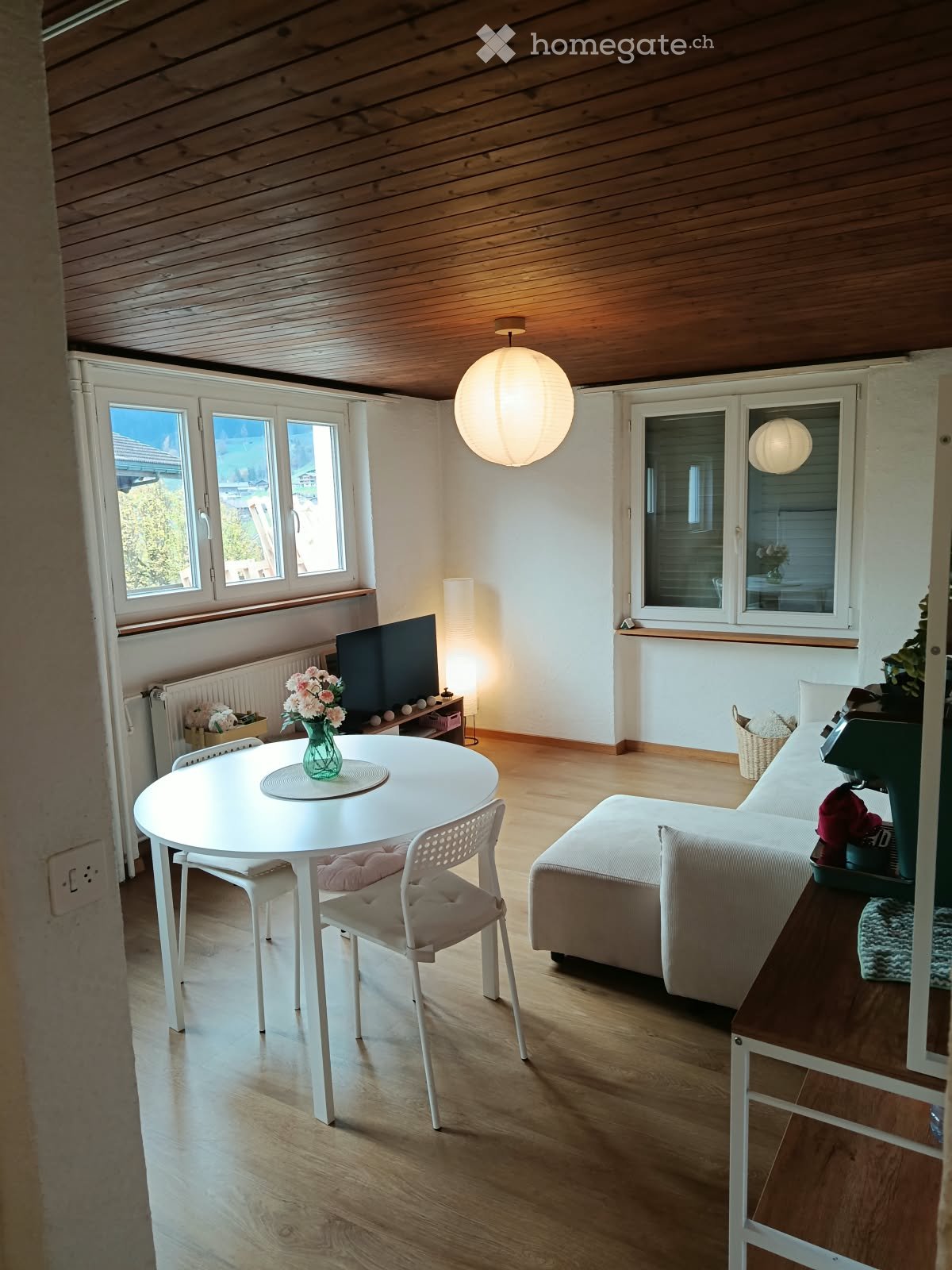 A modern, well-lit living room with wooden flooring, white walls, and large windows. Features include a white sofa, a small round table with chairs, a television, and decorative vases.