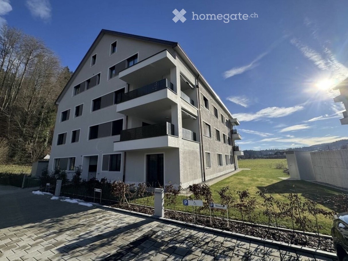 Modern multi-story residential building with balconies, parking area, and landscaped surroundings.