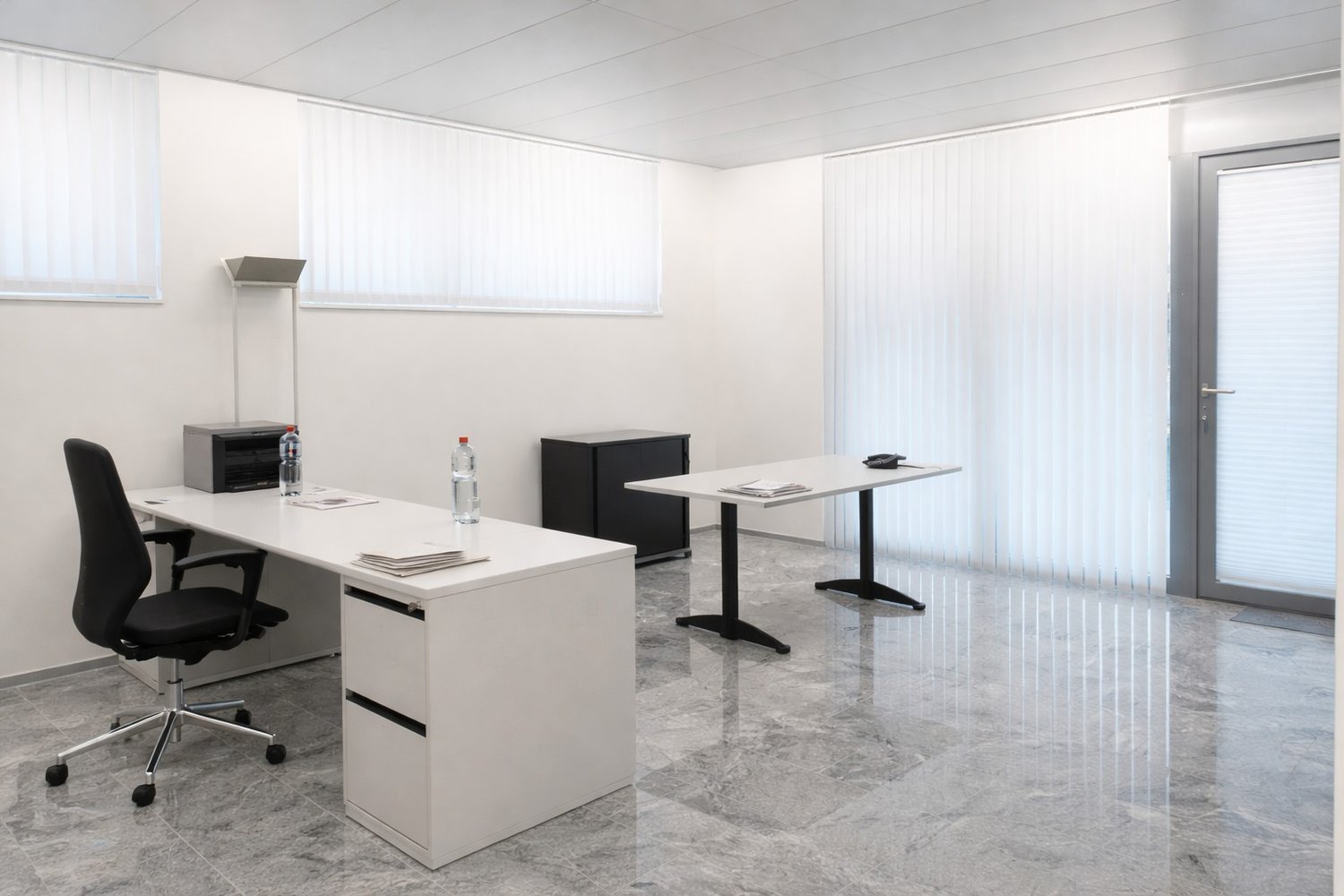 A minimalist office room with white walls and floors, featuring two desks with chairs, a small table, a cabinet, a lamp, and two bottles on the desks.