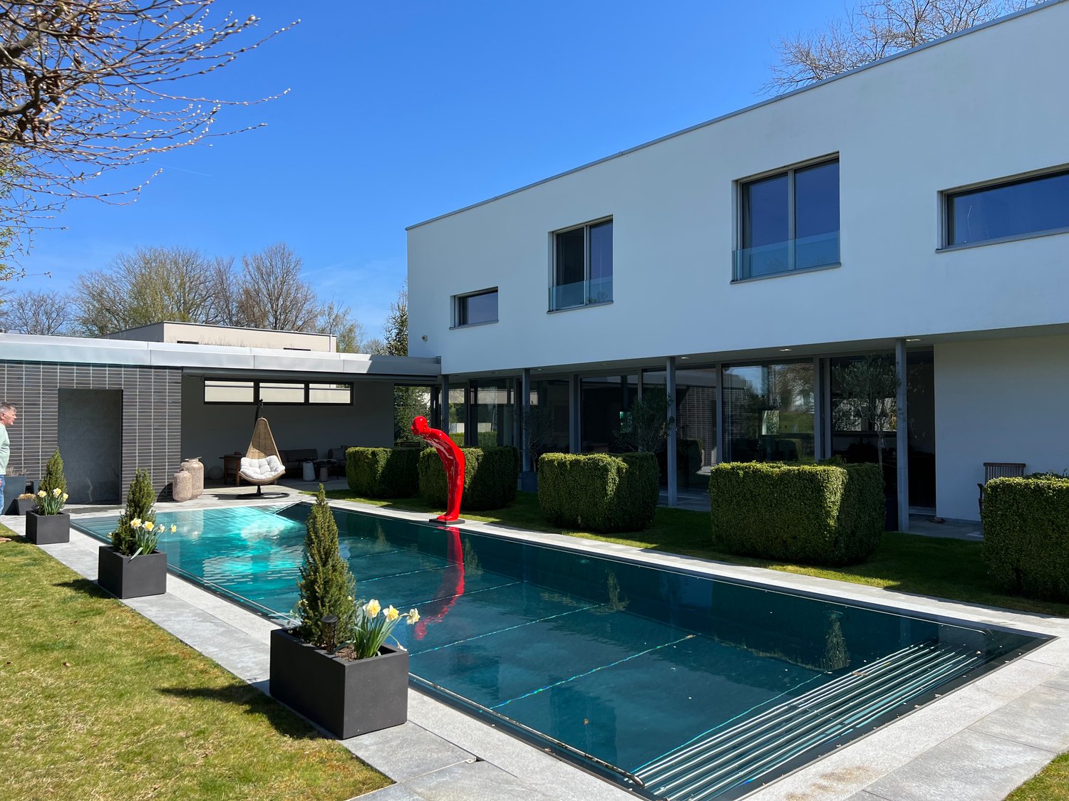 Modern two-story house with a large swimming pool in the backyard. The house has a sleek, minimalist design with large windows and a balcony. The pool is surrounded by landscaped gardens and a red sculpture is placed near the pool.
