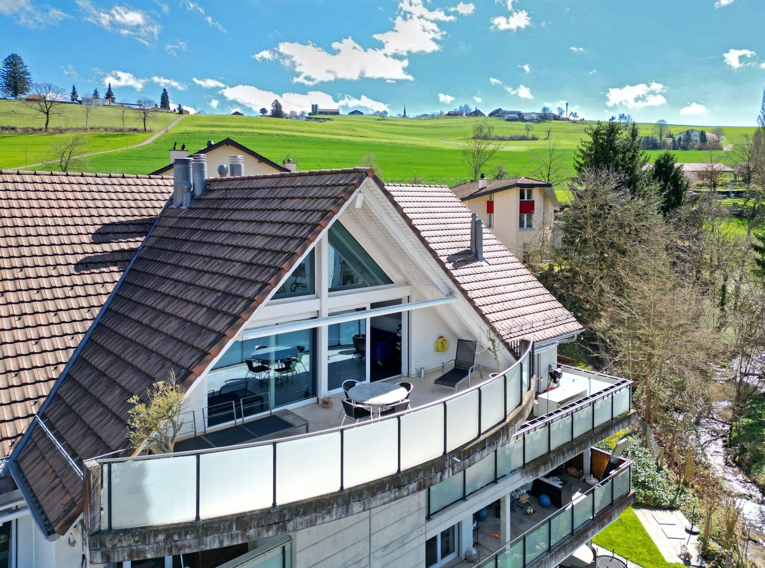 2 story house, brown roof, large windows, balcony with outdoor table and chairs, large lawn, green landscape