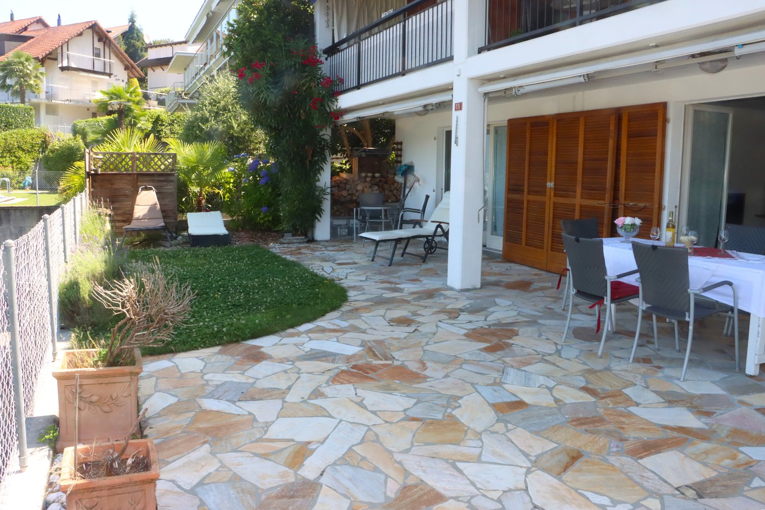 stone floored patio with dining table, chairs, 2 lounge chairs, balcony, shrubs and trees