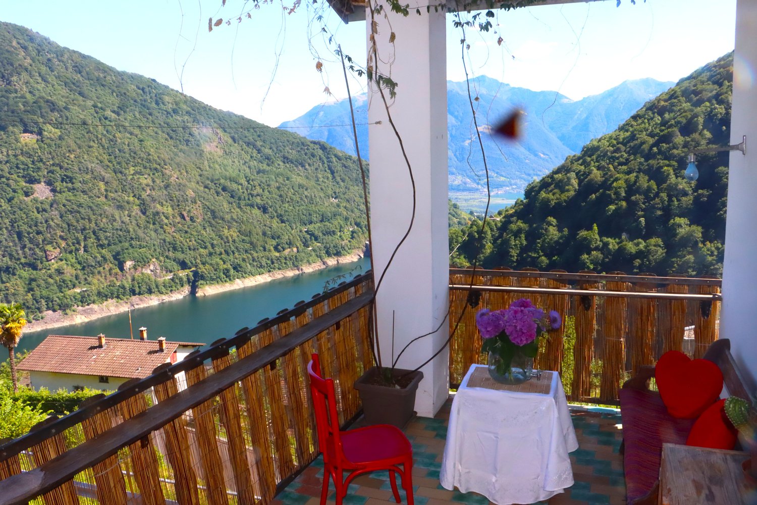 Balcony with wooden railings, red chair, white table with flowers, red pillows, view of mountains and lake