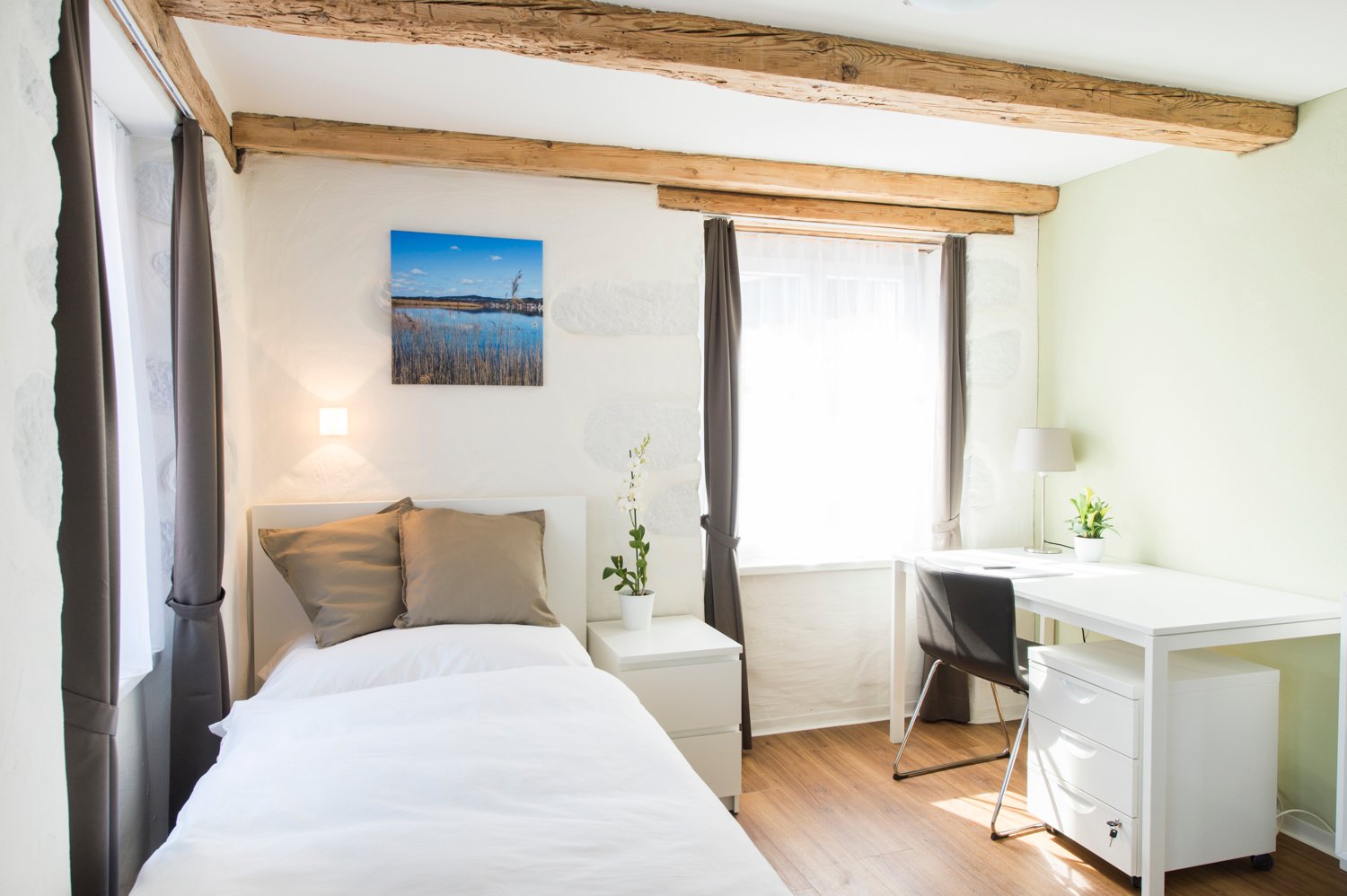 Cozy bedroom with exposed wooden beams, white walls, large window, bed with beige bedding, nightstand, and a desk with a chair.
