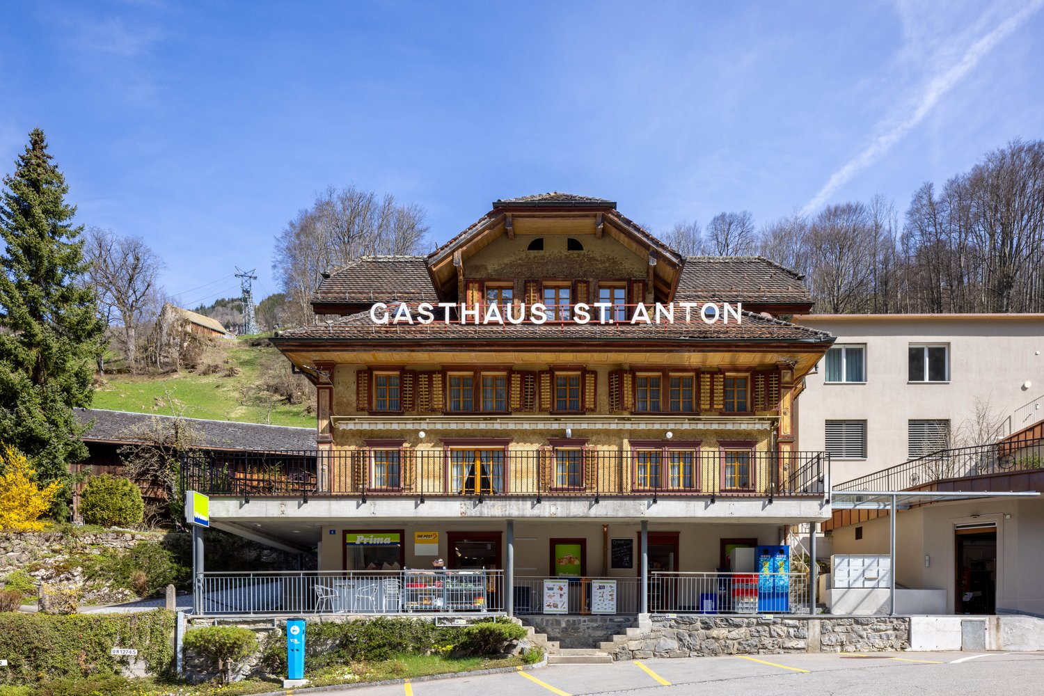A traditional Swiss-style wooden building with a large balcony and terrace, housing a Gasthaus (inn or guesthouse) called 'Gasthaus St. Anton'. The building has a prominent sign displaying the name and is surrounded by trees and a grassy area.