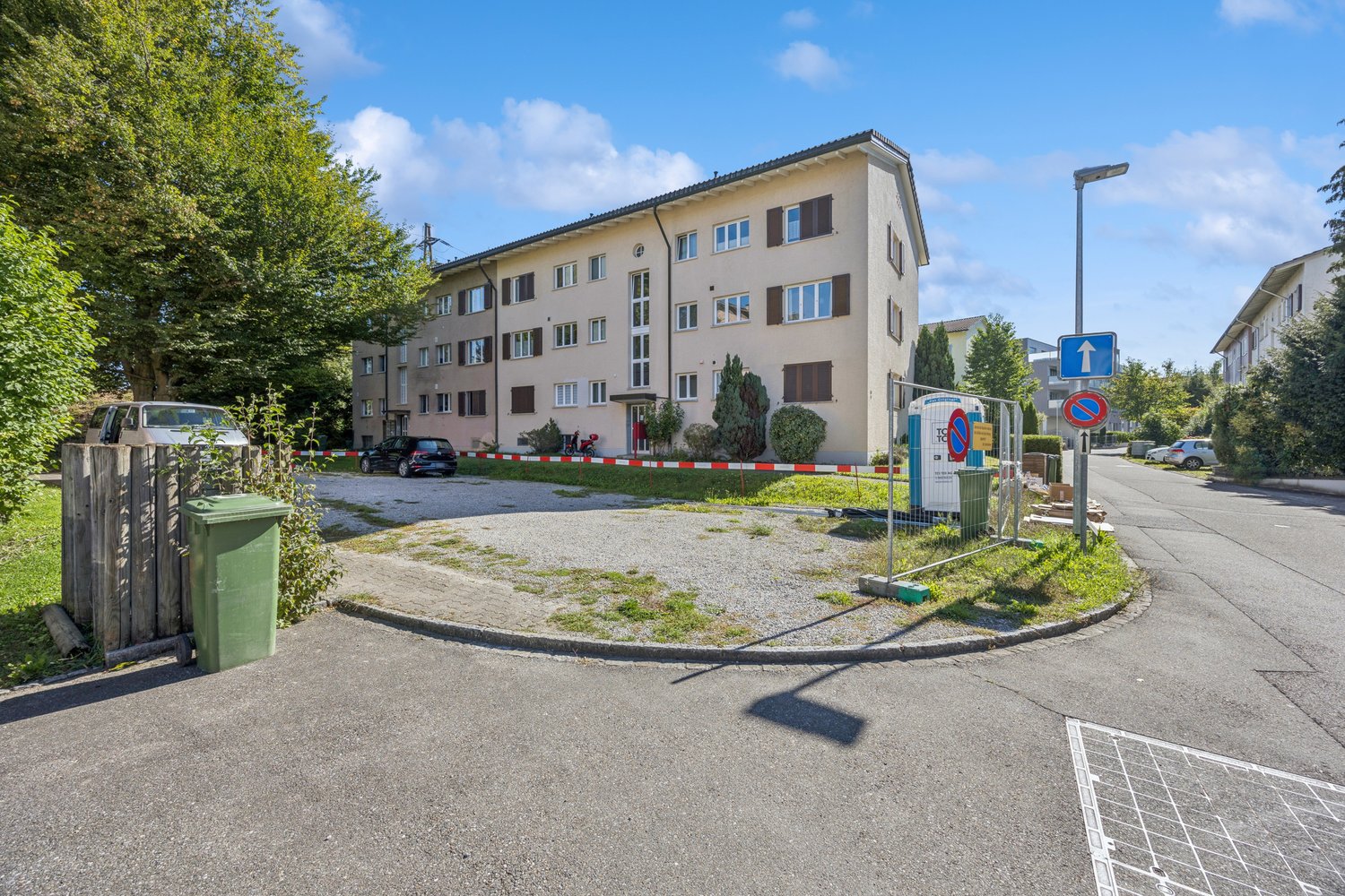 3 story building, beige exterior, multiple windows, cars parked in front, trees, street light, trash bin