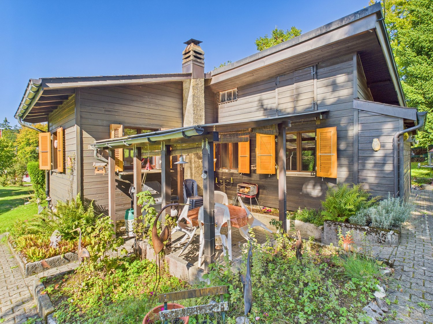 wood house, orange shutters, garden with plants, brick pathway, chimney, porch with table and chairs, grill, trees in the background