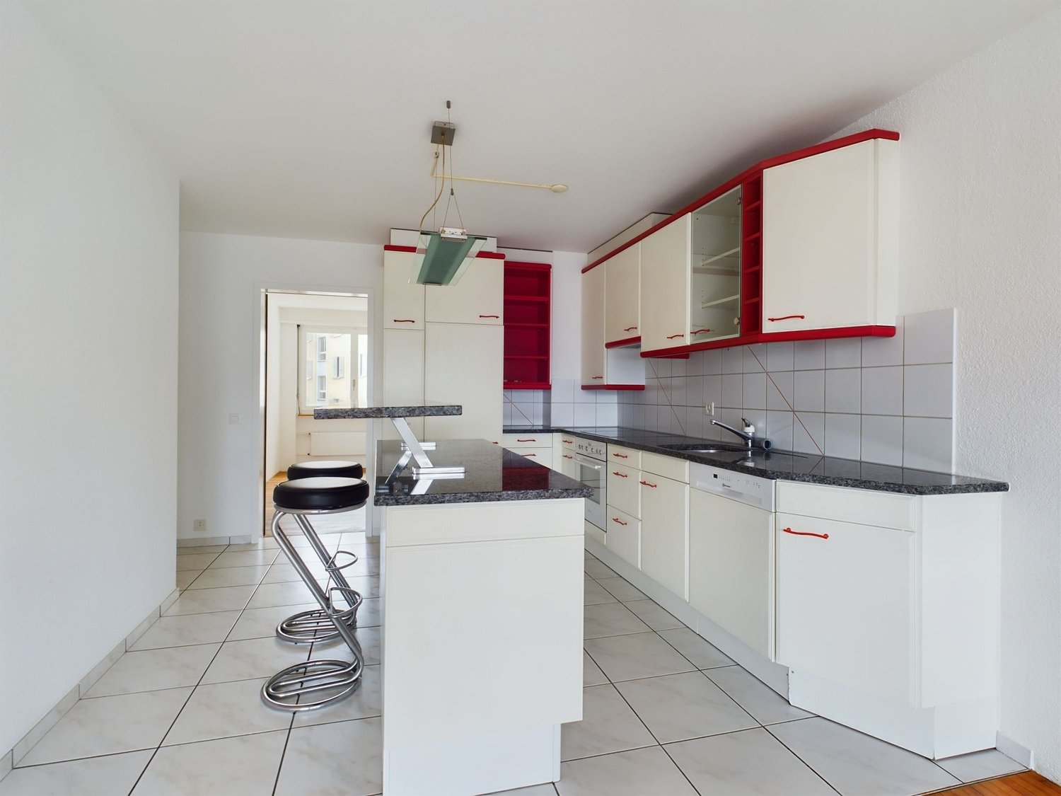 Spacious kitchen with white cabinets, red accents, granite countertops, tiled flooring, and a dishwasher. The kitchen is open to a dining area with a bar counter and stools.
