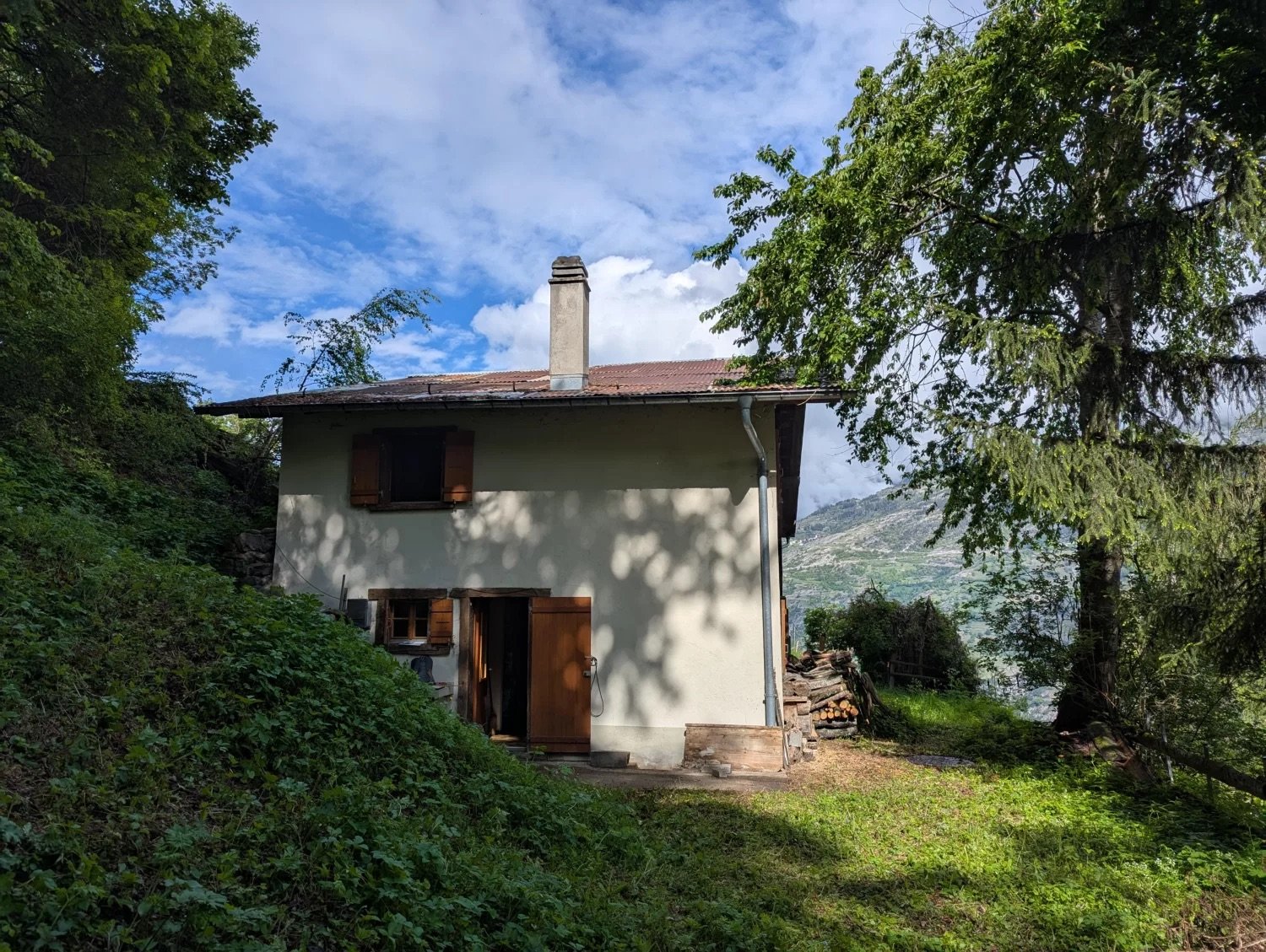 Two-story house with a chimney, surrounded by lush greenery and trees, with a balcony or terrace visible.