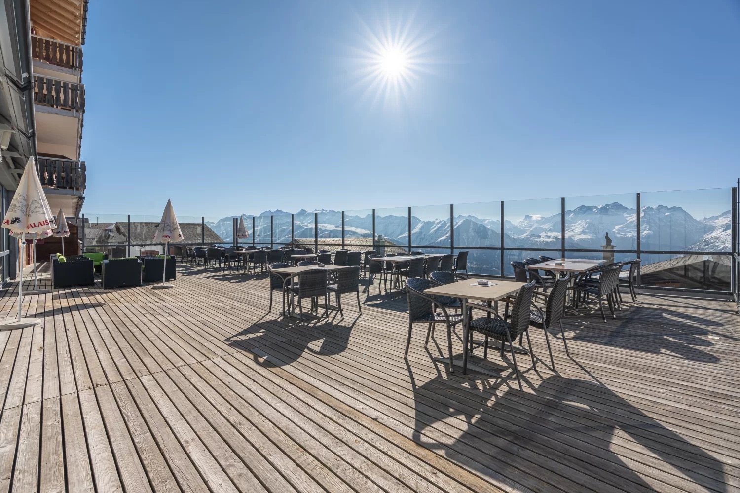 Outdoor dining area with wooden deck, tables and chairs, surrounded by glass railings offering a panoramic view of snow-capped mountains under a bright, sunny sky
