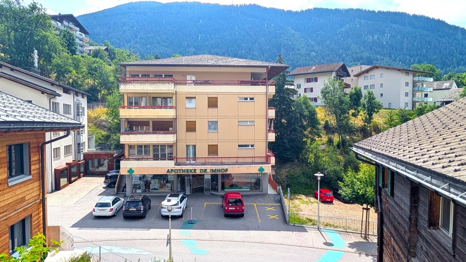 A multi-story residential building with a pharmacy on the ground floor, surrounded by trees and mountains in the background. The building has balconies and there are several cars parked in the lot in front of the building.