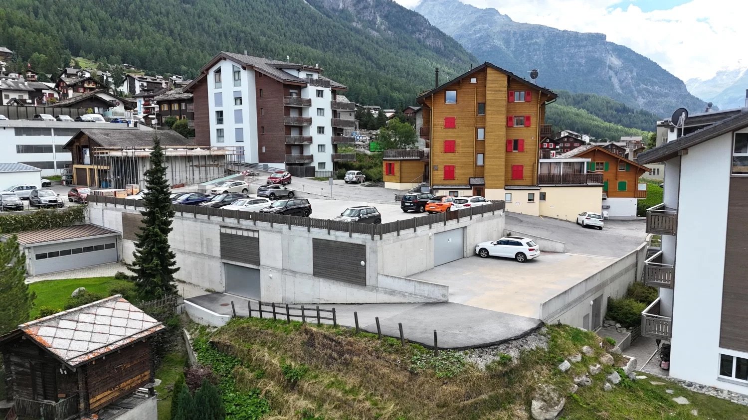 The image shows a residential area in a mountainous region. There are several multi-story apartment buildings and houses, some with colorful facades. There is a parking lot in the foreground with several cars parked. The surrounding landscape includes for