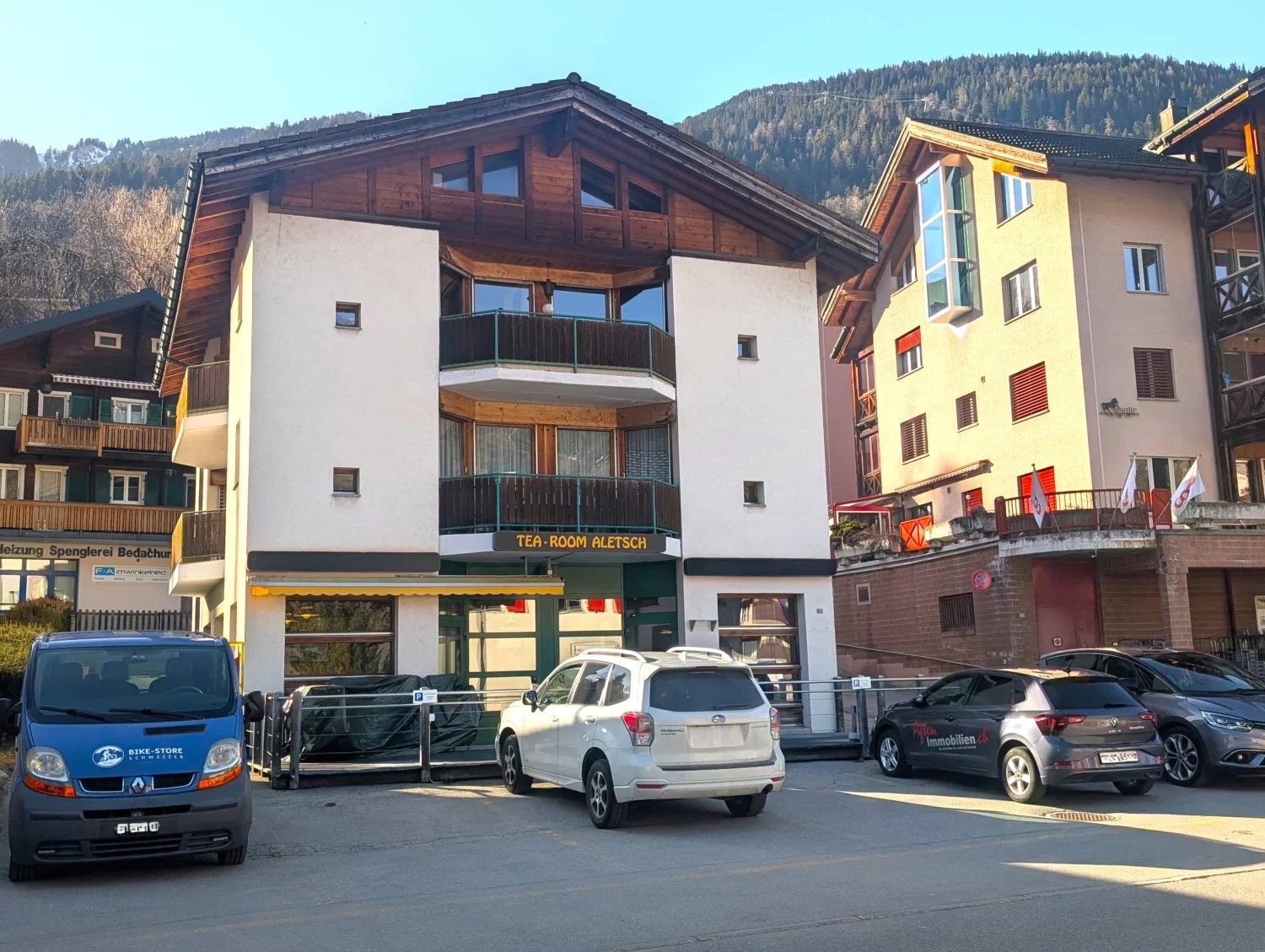 A three-story building with white and brown exterior, balconies on the second floor, and a name board 'TEA-ROOM ALETSCH'. There are several cars parked in front.