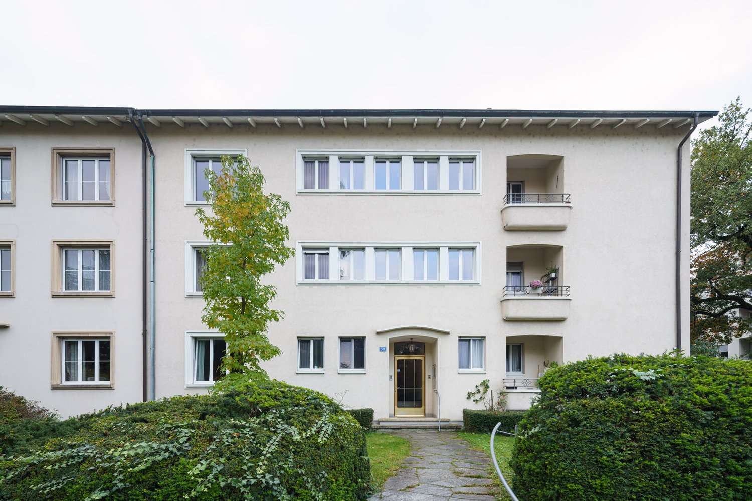 3 story building, white exterior, multiple windows, balconies, entrance with door and steps, hedges and tree in front