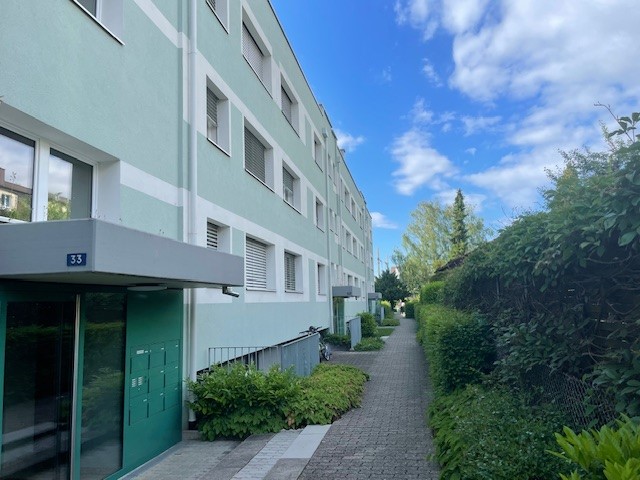 A large building with a pathway in front of it, green door, green awning, white and green walls, bicycle parked on the side.