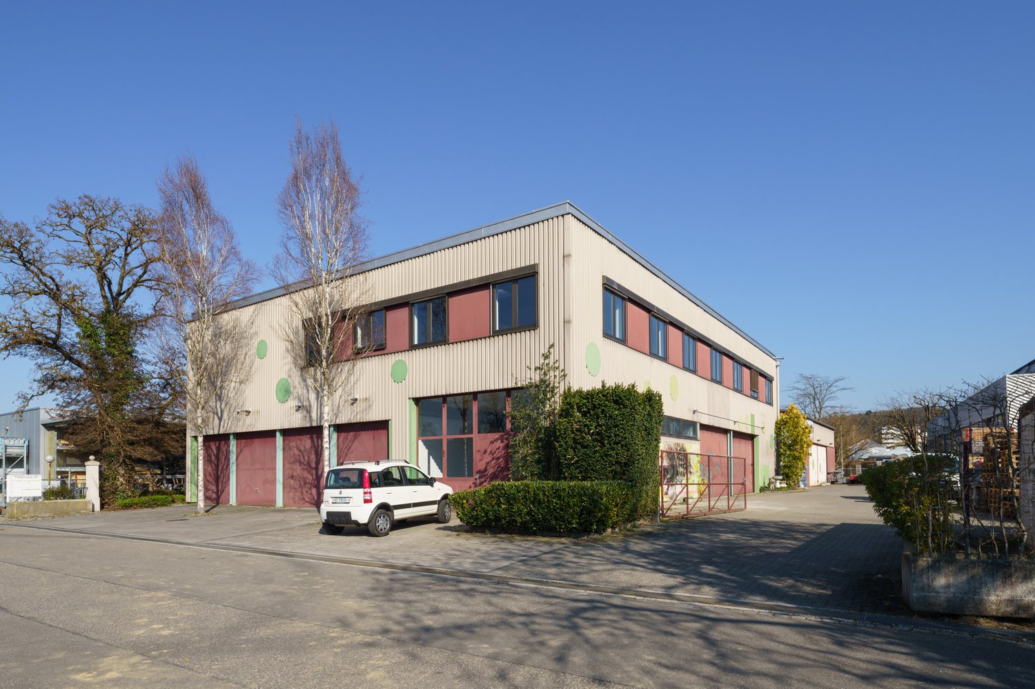 Industrial building with red garage doors, white car parked in front, large windows, green bushes, clear sky