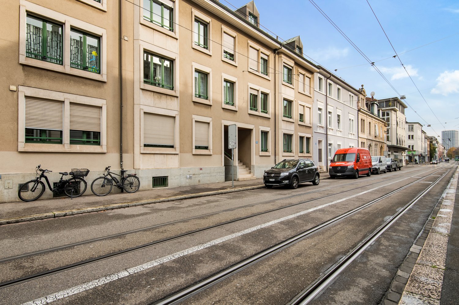 Beige building, many windows, bicycles parked in front, street, tram tracks, cars parked on the road, red van.